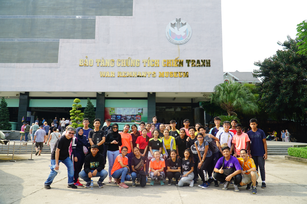 A large group of students and adults posing in front of the War Remnants Museum in Vietnam.